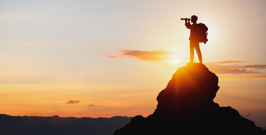 Silhouette of man holding binoculars on mountain peak against bright sunlight sky background. growth leadership career