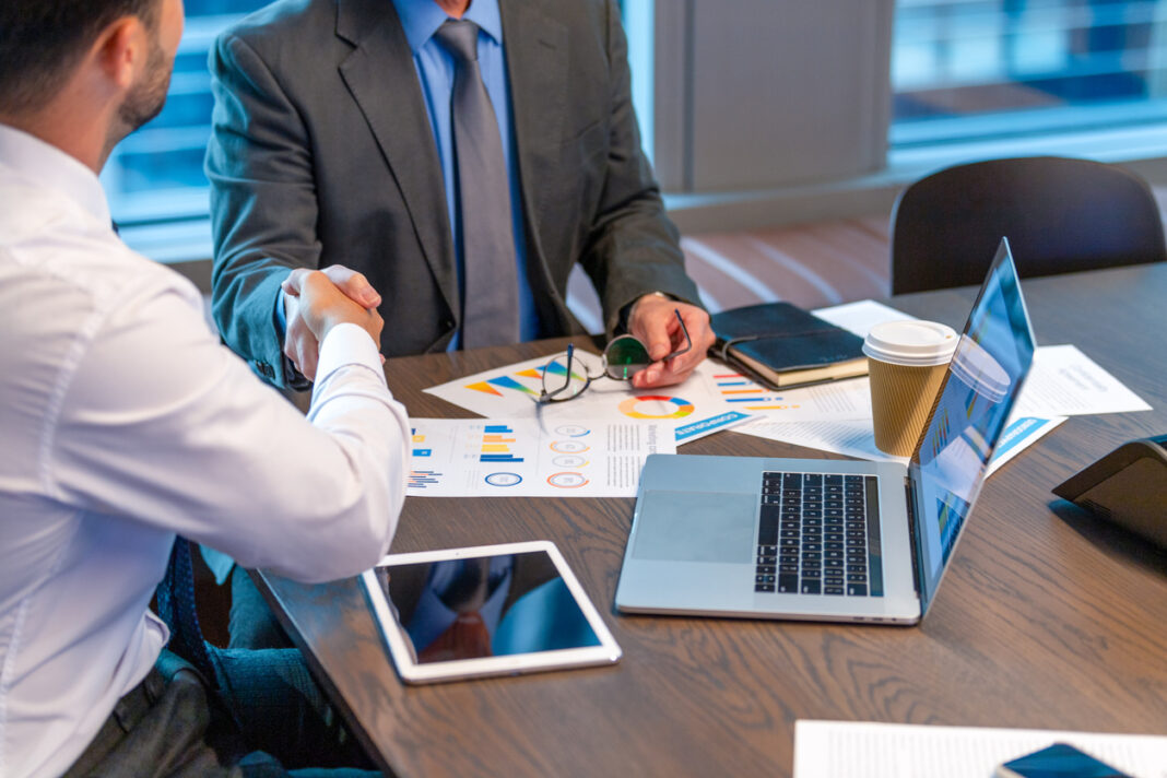 Businessmen shaking hands. Both are dressed in business clothing and there is paperwork with finance graphs and charts